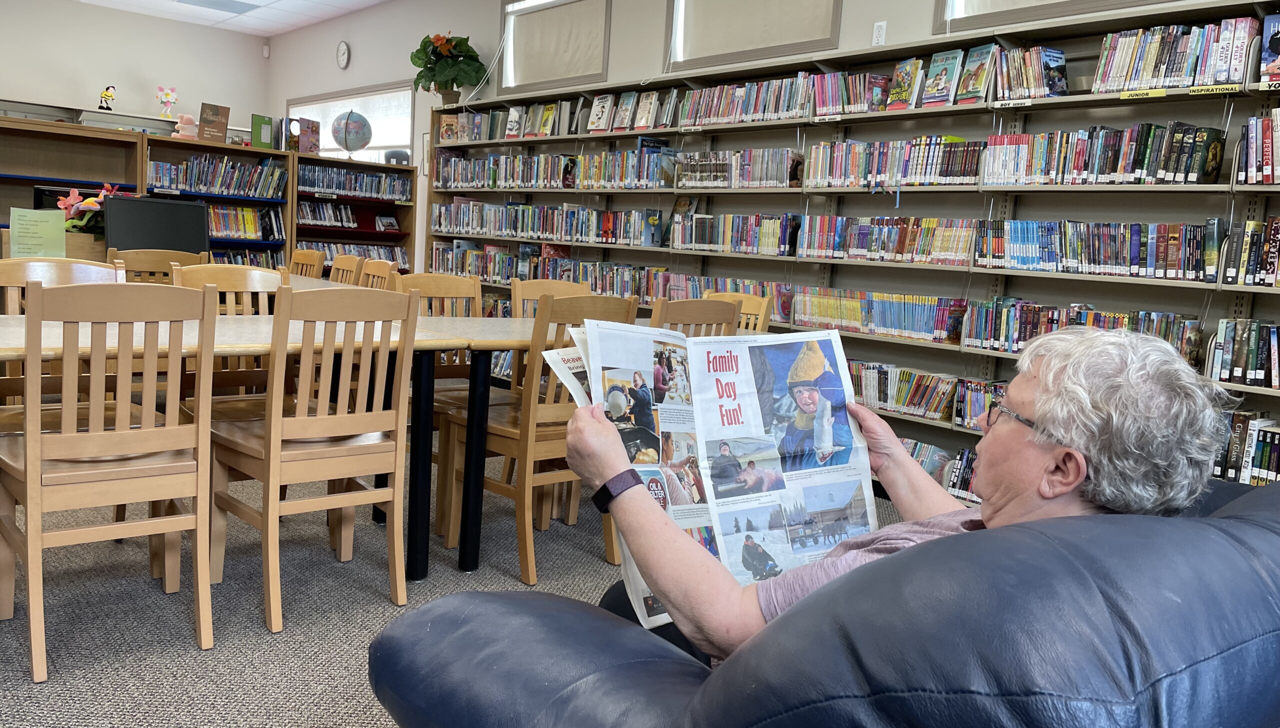 Interior shot of a person sitting in a chair reading a newspaper in front of bookshelves.