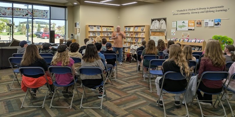 Interior shot of Penhold Library. Group of people sitting on chairs facing away from the camera, towards a man standing and giving a presentation.